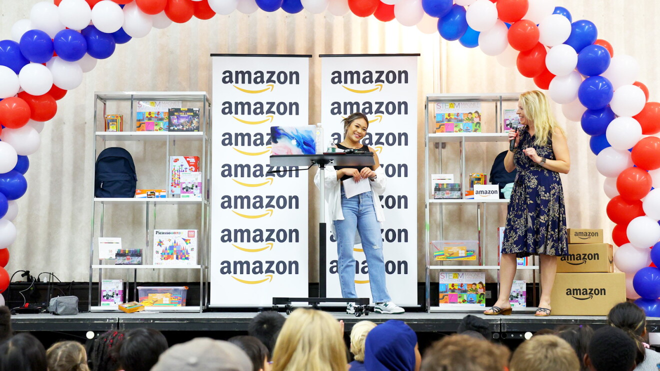 Suni Lee stands next to the principal on a stage in front of a group of students at an elementary school. There is back-to-school items and an Amazon sign in the background.