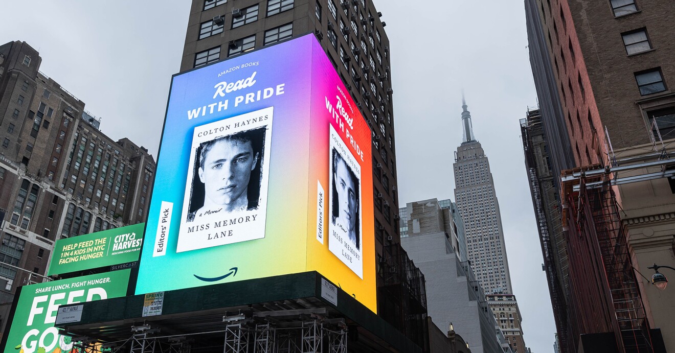 An image of a Times Square billboard with a book cover on it for "Miss Memory Lane." The text above the cover says "Read with Pride," and there is a tag on the side of the cover that says "Editor's pick." There is a rainbow-colored background behind the book and the Amazon smile logo below.