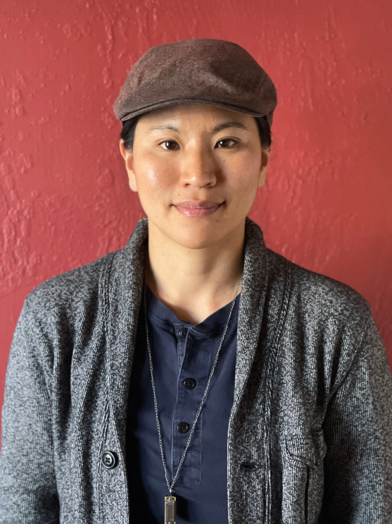 A woman wearing a newsboy cap, henley shirt, sweater, and necklace sits in front of a red wall. 