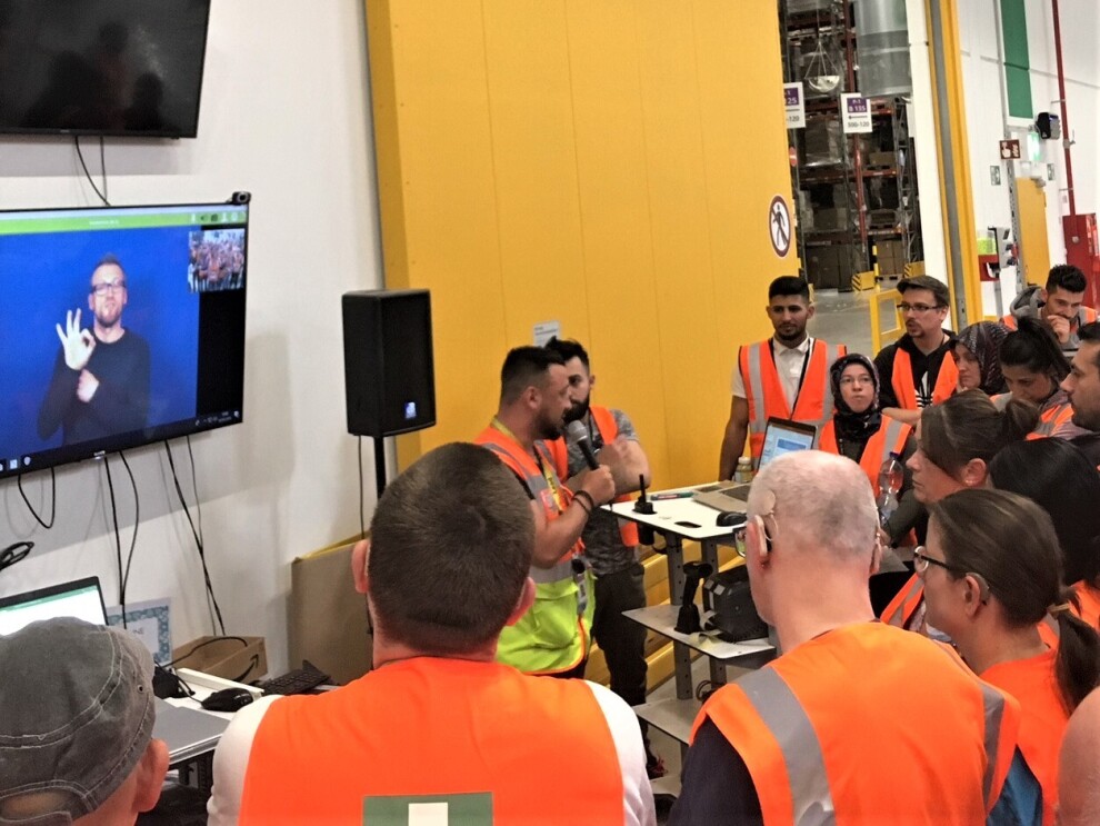 Das Bild zeigt Mitarbeiter bei Start-Meeting: Ein Manager spricht in eine Mikrofon, an der Wand hängt ein Screen, auf dem gerade eine Videokonferenz mit einem Gebärdendolmetscher läuft.
