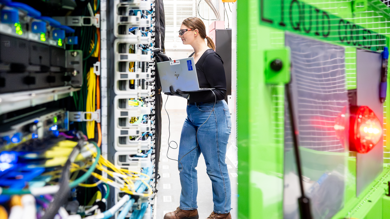 Technician servicing ambiguous rack with laptop, near "Liquid cooled only" sign.