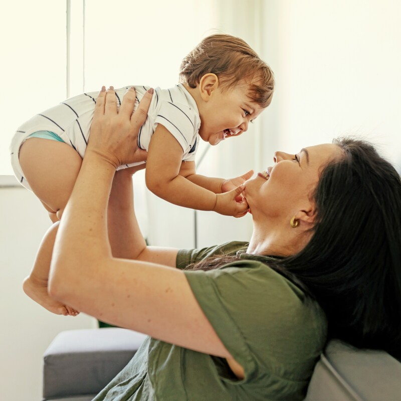 Mãe, sentada em um sofá, levanta bebê. Eles sorriem