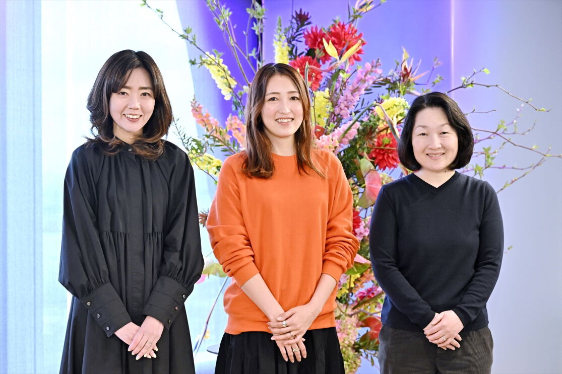 黒いドレスを着ている女性、オレンジ色のセーターを着ている女性、そして黒いTシャツを着ている女性が大きな花装飾の前に立っている。