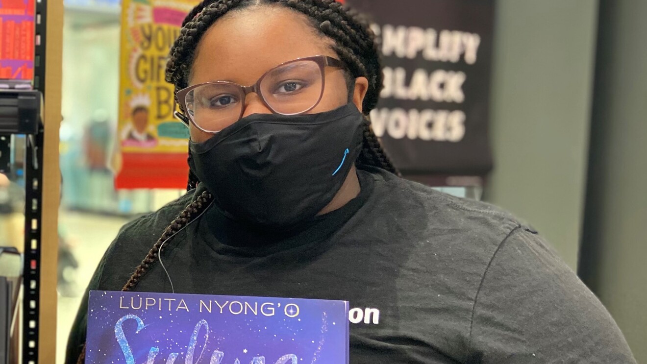 An image of a woman standing in an Amazon bookstore smiling for a photo under her mask. She is holding a children's book titled "Sulwe," authored by Lupita Nyong'o.