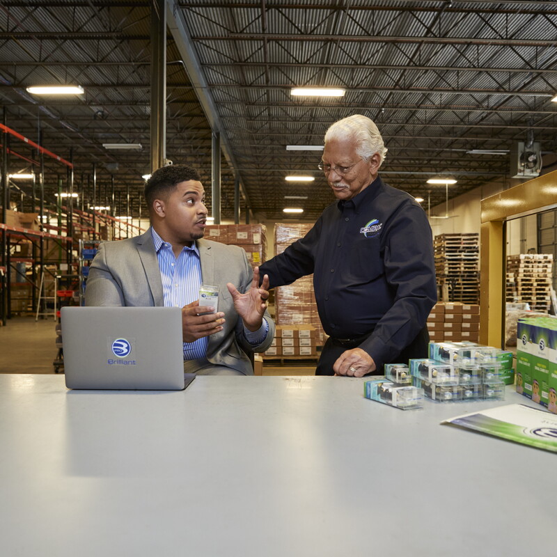 Two men speak to one another at a tabletop surface in a warehouse facility