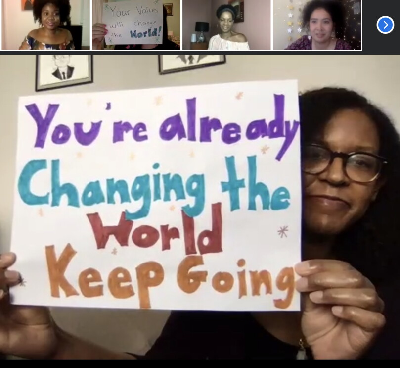 An image of a woman on a video call holding up a hand-drawn sign to her camera that says "You're already changing the world. Keep going." There are women on the call with her and their faces are in the smaller boxes at the top of the screen.