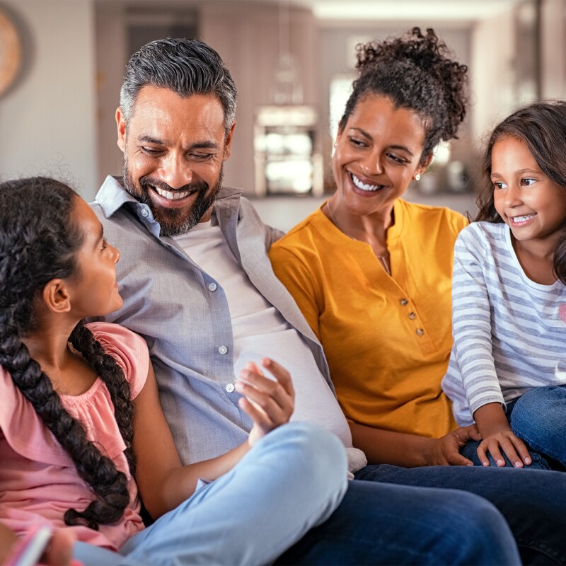 Uma família, sentada em um sofá, conversando e sorrindo. São duas crianças, um homem e uma mulher