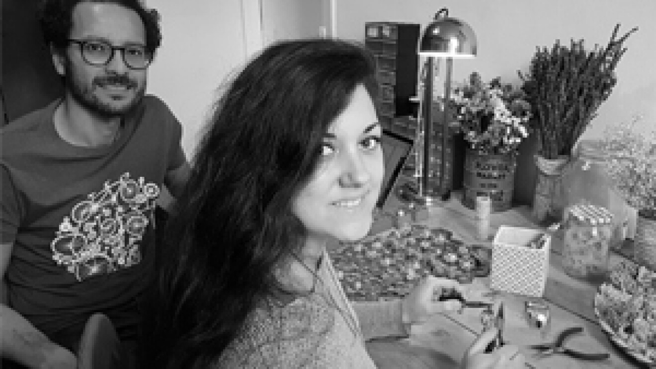 Les deux fondateurs de Trois Fenêtres se tiennent devant une table où est posé du matériel de création artisanale ainsi qu'une lampe et des bouquets de fleurs. C'est une photo noir et blanc.