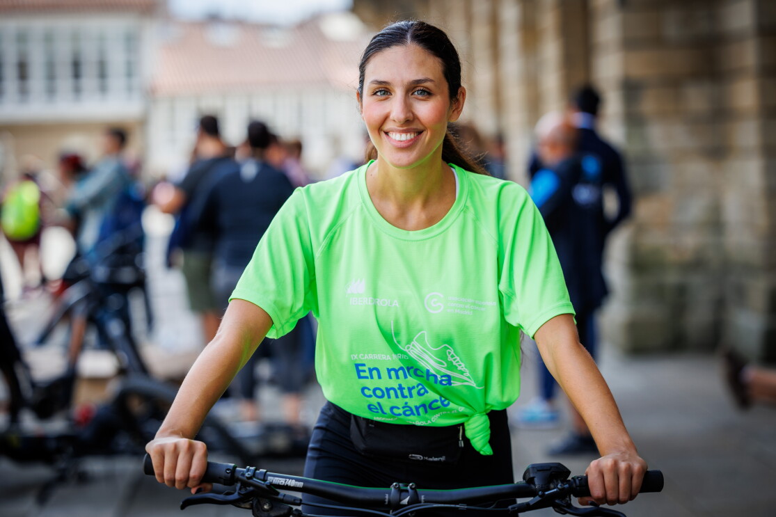 Laura Cano, trabajadora del centro logístico de Illescas. En la plaza del Obradoiro de Santiago de Compostela está Laura encima de una bicicleta . Apoya las manos en el manillar . Va con una camiseta de color verde que pone En marcha contra el cáncer. Lleva el pelo recogido con una coleta. Tiene los ojos azules y está sonriendo. 