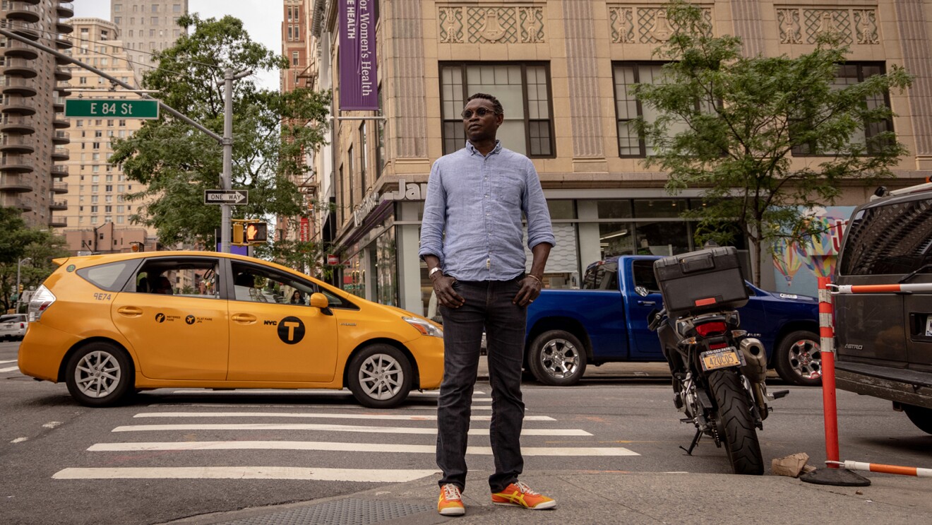 An image of a man standing on the sidewalk in the middle of a large city. There are cars and a taxi cab on the street behind him.