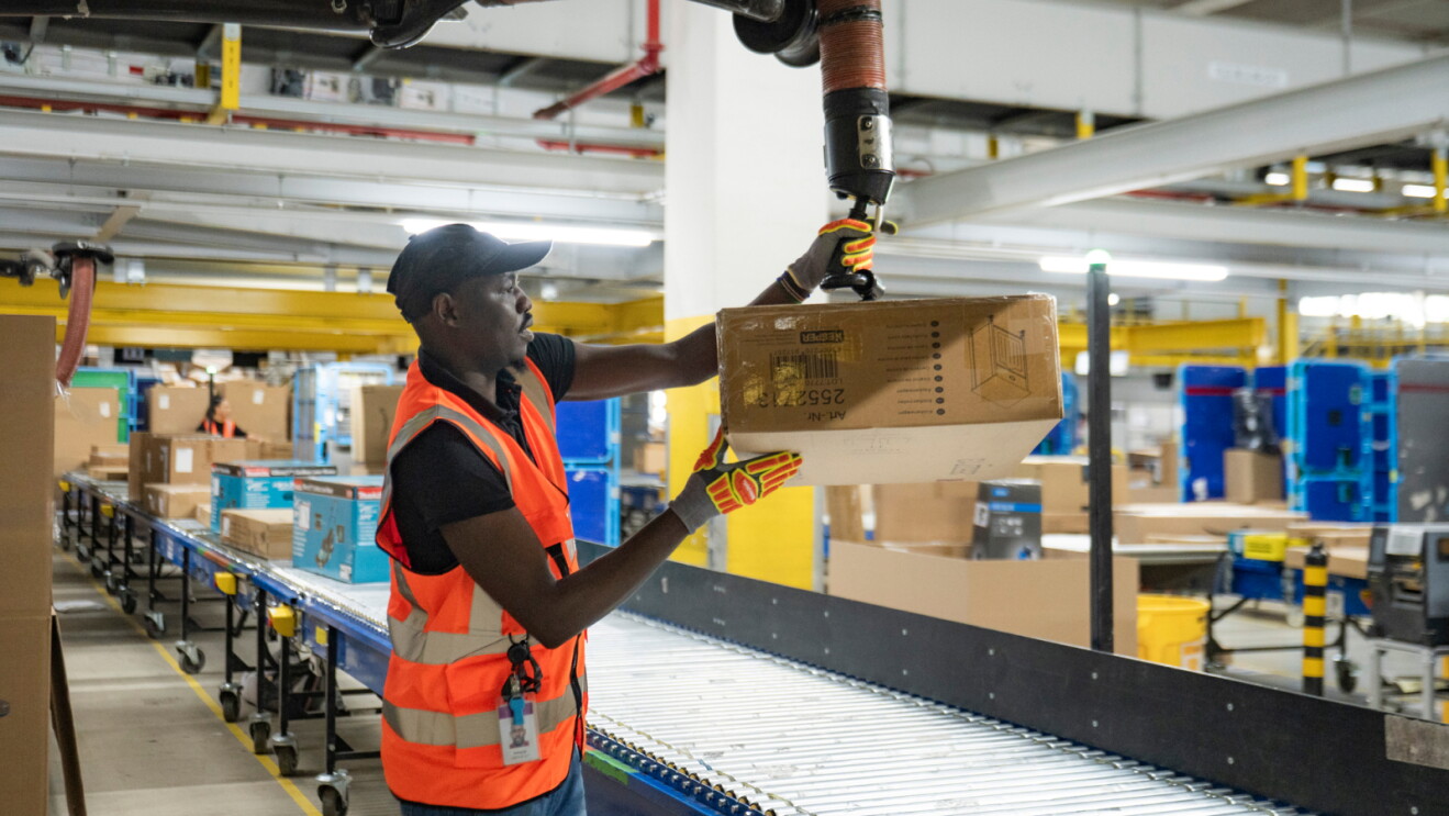 A man wearing a cap, an orange high-visibility vest and protective gloves handles a cardboard box beside a conveyor belt in a fulfilment centre. Boxes, equipment and a colleague are visible in the background.