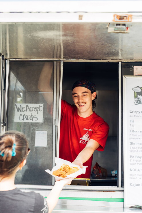 A man wearing a red t-shirt leans out of a food truck window, handing a basket of chicken strips and fries to a woman standing outside the truck.