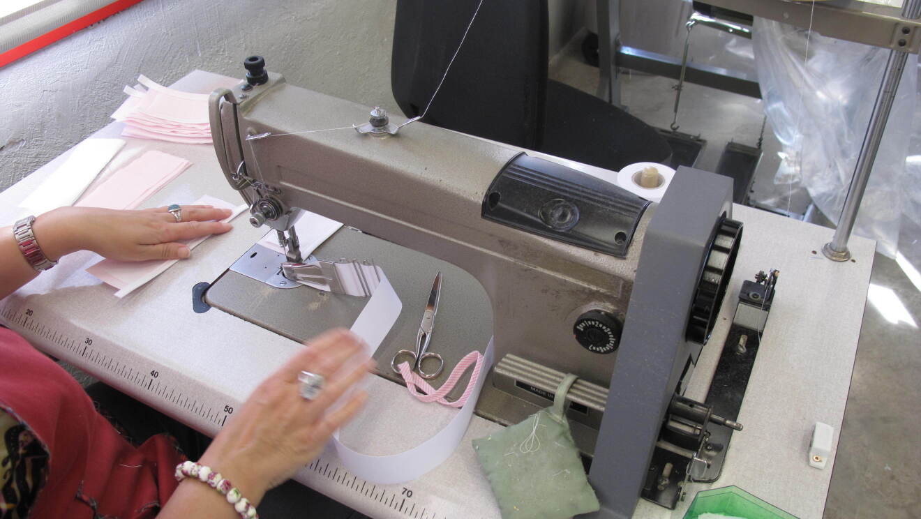 Plano picado de una mujer cosiendo las asas de una canastilla de bebé. Las asas son de color blanco. La mujer lleva una bata de color rojo y en la mano derecha lleva una pulsera y un azul, y en la mano izquierda el reloj y otro anillo. La máquina de coser es de color gris. 