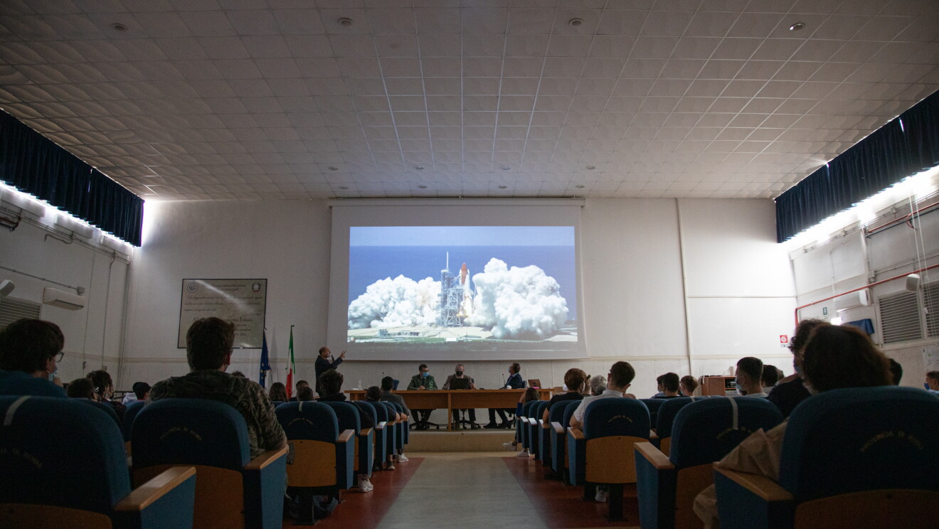 Sala riunione all'interno del magazzino di Colleferro di Amazon. In fondo, in mezzo alla platea di sedie è appeso sul muro un telo per proiettore sul quale si vede l'immagine di un razzo spaziale in procinto di decollare. In platea, un gruppo di studenti ripresi di spalle