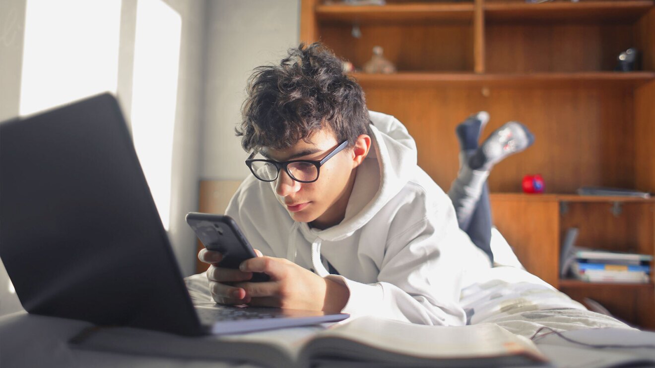 A teenage boy lays on his bed while using a cell phone and a laptop. 