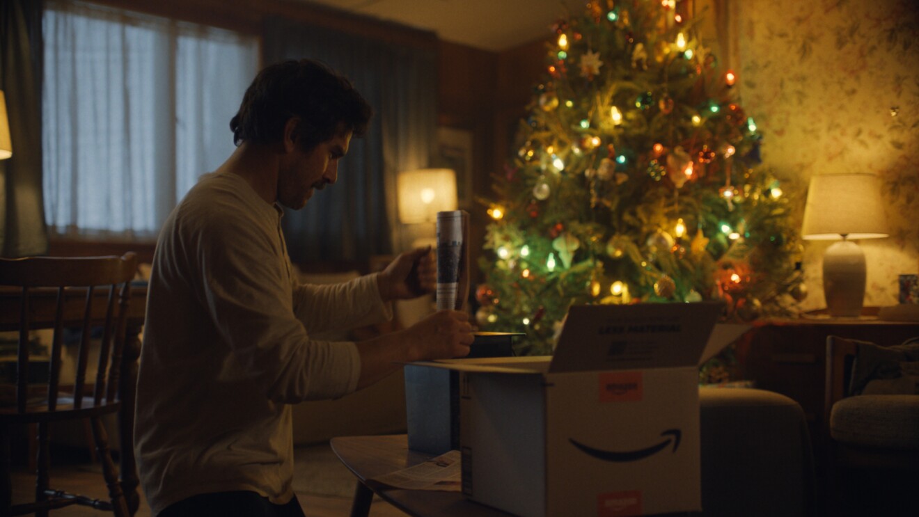 Dentro del salón de una casa con decoración de los años 60, con papel estampado en la pared, mesa y sillas de madera y cortinas oscuras, hay dos lámparas de mesa y un árbol de Navidad decorado y con luces de colores. En primer plano hay un señor vestido con un jersey de color blanco, abriendo una caja de cartón de Amazon con una sonrisa. El señor lleva el pelo corto y es moreno y lleva bigote. 