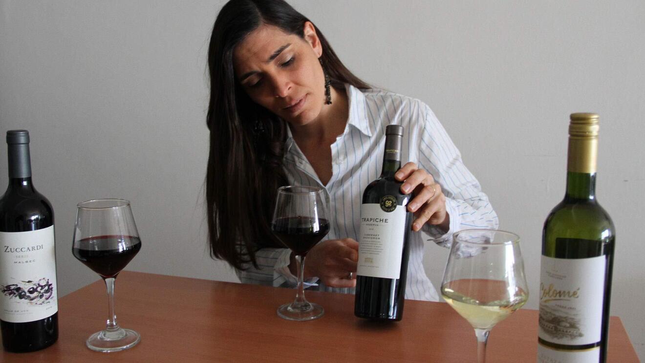 A woman sits at a table with three wine glasses of various colors and two bottles for a virtual wine tasting.
