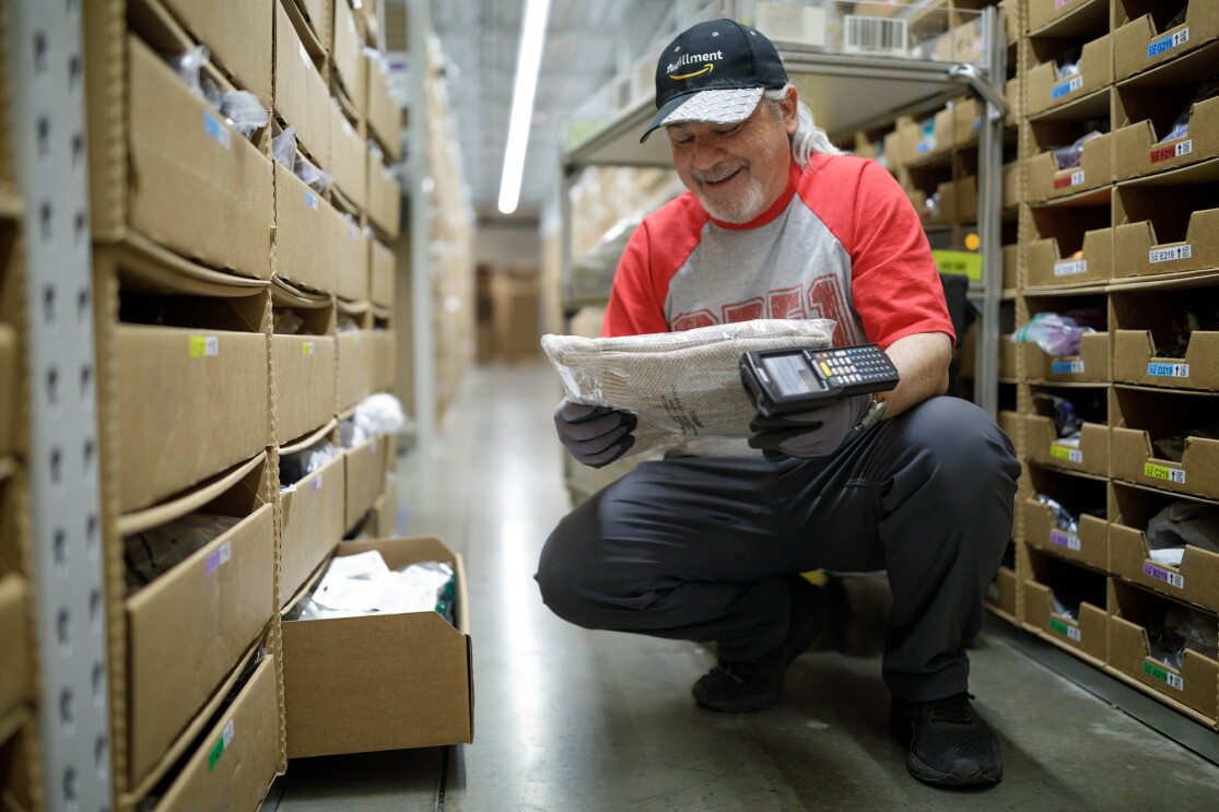 A man in a baseball cap with the Amazon logo smiles. He holds a scanning device in one hand and a piece of item of clothing in the other. To his side and in the background, there are metal shelving units with cardboard bins. 