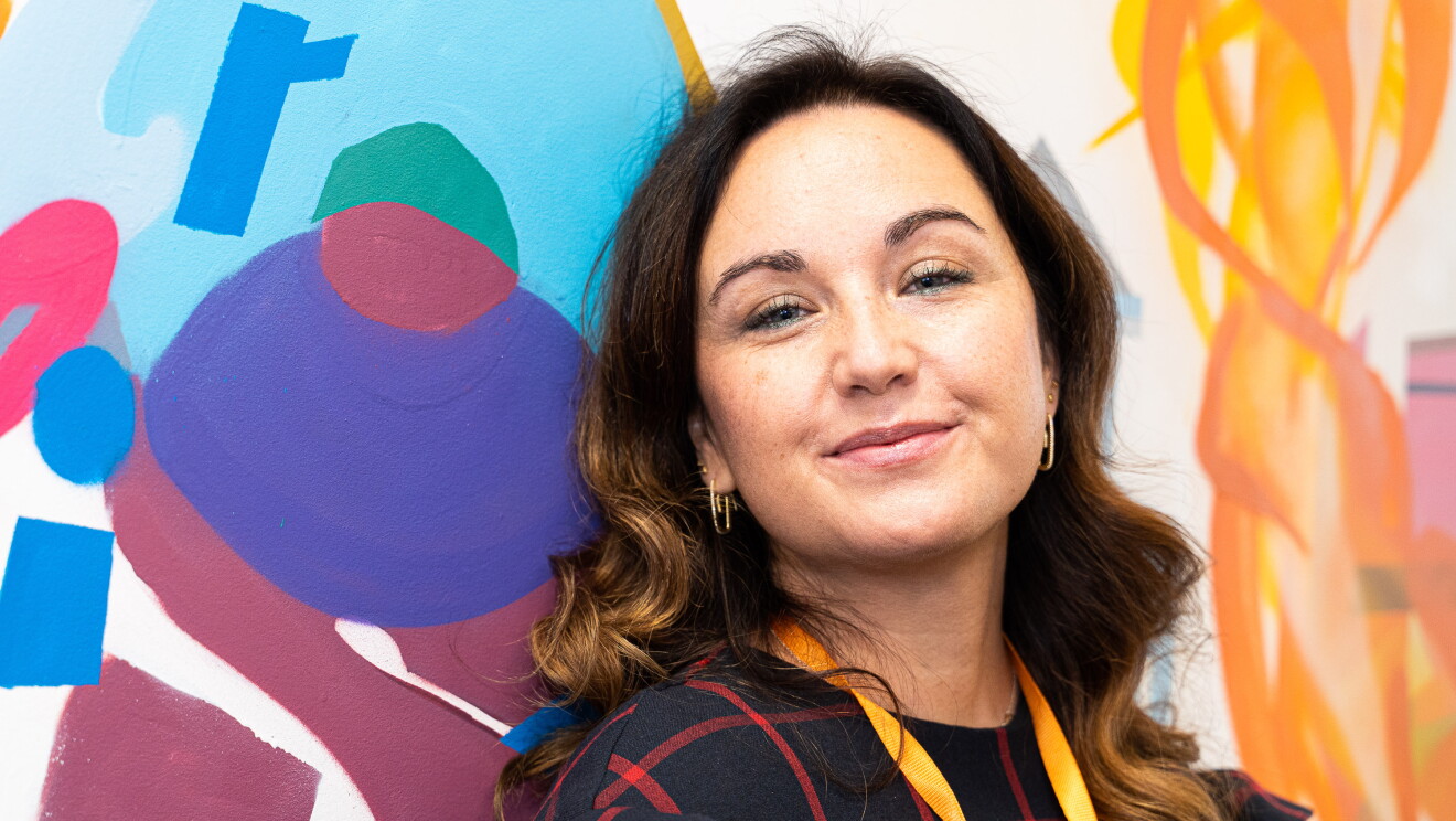 Elena Bris trabajando en las oficinas de Amazon en Madrid.  Elena Bris trabaja en un proyecto regulatorio de mercancías peligrosas. Mirando a cámara en primer plano. De fondo una prared estampada con pinceladas de difernetes colores. Elena lleva un bagde con una cinta de color amarillo y un vestido azul con cuadros de color rojo. Tiene el pelo largo y ondulador. Lleva pendientes y los ojos pintados de azull. Lleva pendientes. 