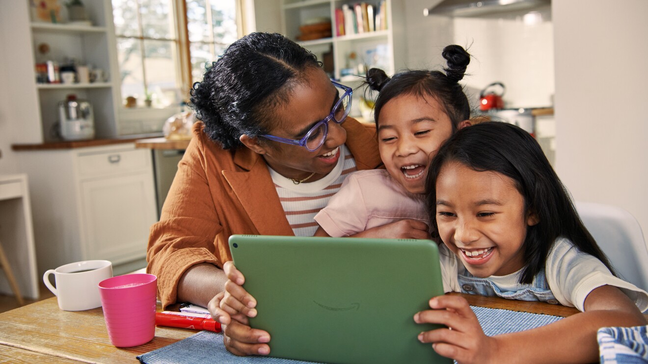 Mother and two daughters laugh at kitchen table while viewing content on an Amazon tablet. 
