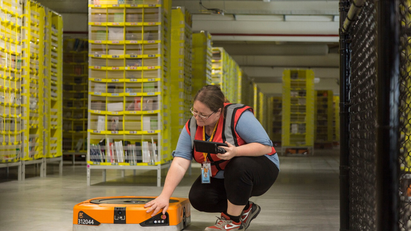 Foto di Maria Stella Carnà che regge un tablet di controllo e si china su uno dei robot di cui è responsabile all'interno del Centro di Distribuzione Amazon di Passo Corese
