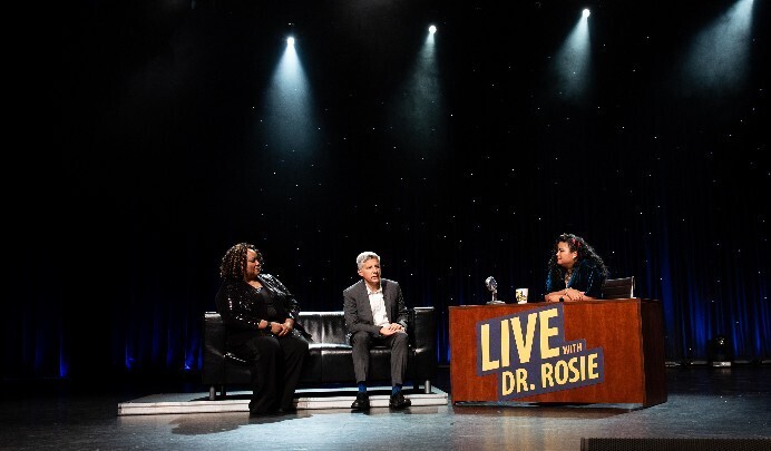 The set of the show, Live with Dr. Rosie. Two people sit on a couch and to their right is Dr. Rosie sitting behind a desk with the logo of the name of the show on the front of the desk.