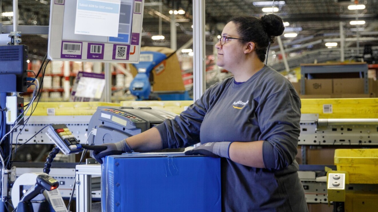 Pracowniczka Amazon w koszulce z logiem, wpatruje się w ekran, naciskając przycisk na klawiaturze, w tle wnętrze centrum logistyki.