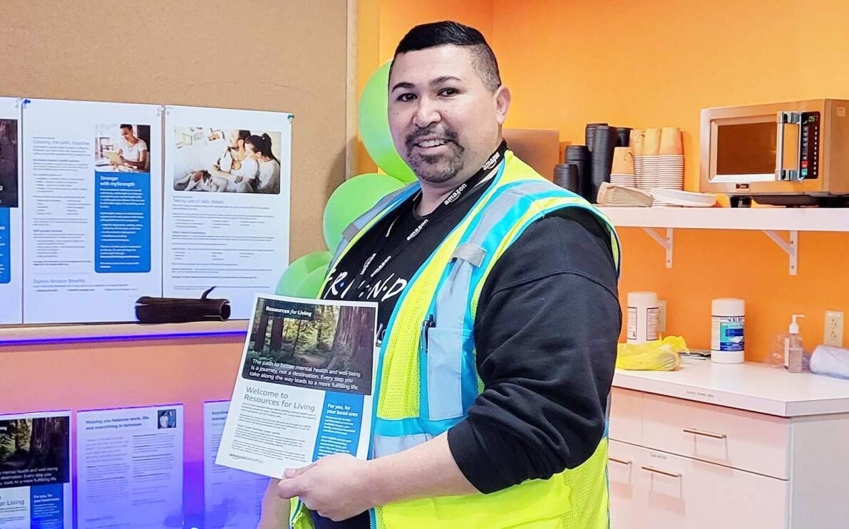 Amazon employee Ernan stands in an employee lounge area holding up a flyer and smiling at the camera. 