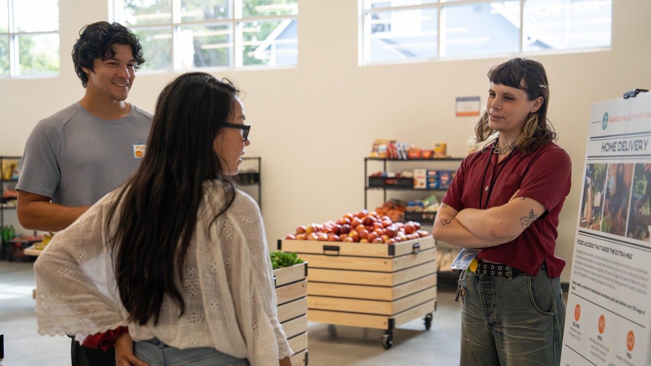 Three people conversing in a food bank with produce and shelves visible