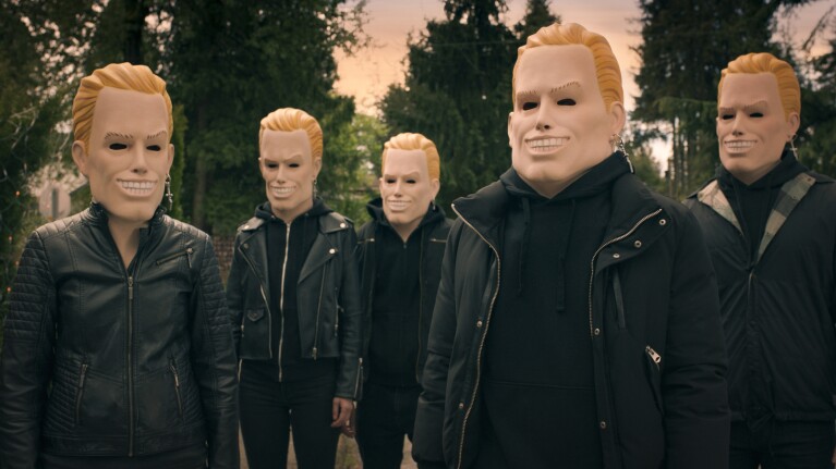 Five figures in black jackets wearing identical blonde masks with wide grins