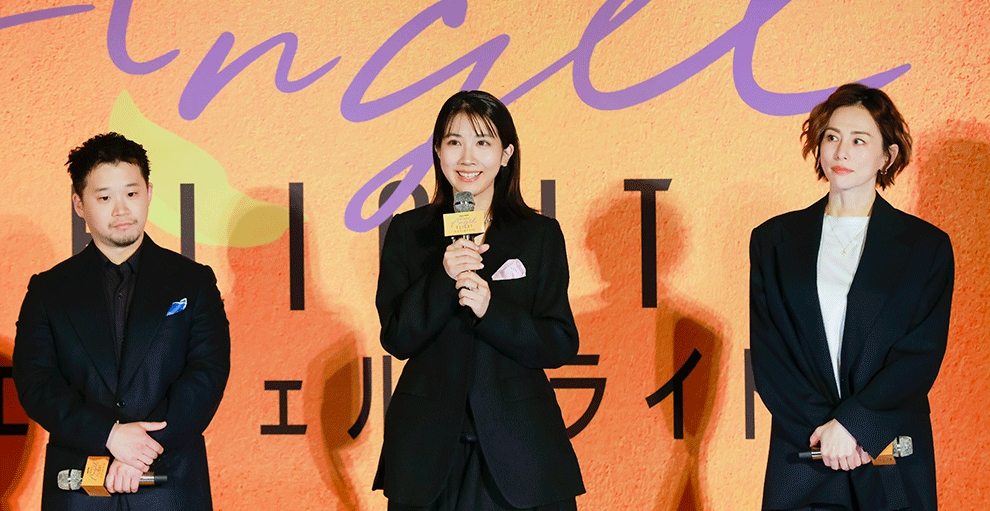 オレンジ色の壁の前、オレンジのステージの上に、黒い服を着て横一列に並んだ3人の人物。全員マイクを手に持ち、中央の人物がマイクで話している。