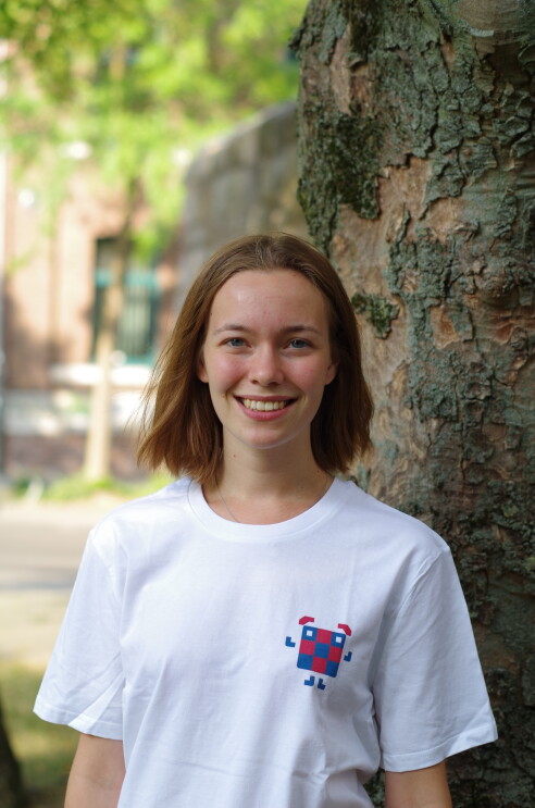 Ein Mädchen mit hellbraunen Haaren und weißem T-shirt steht vor einem Baum und lächelt in die Kamera.
