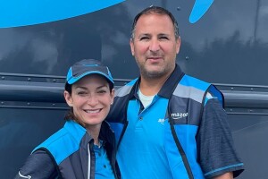 Alison Gatto and her husband wear blue Amazon uniforms and pose in front of a delivery vehicle.