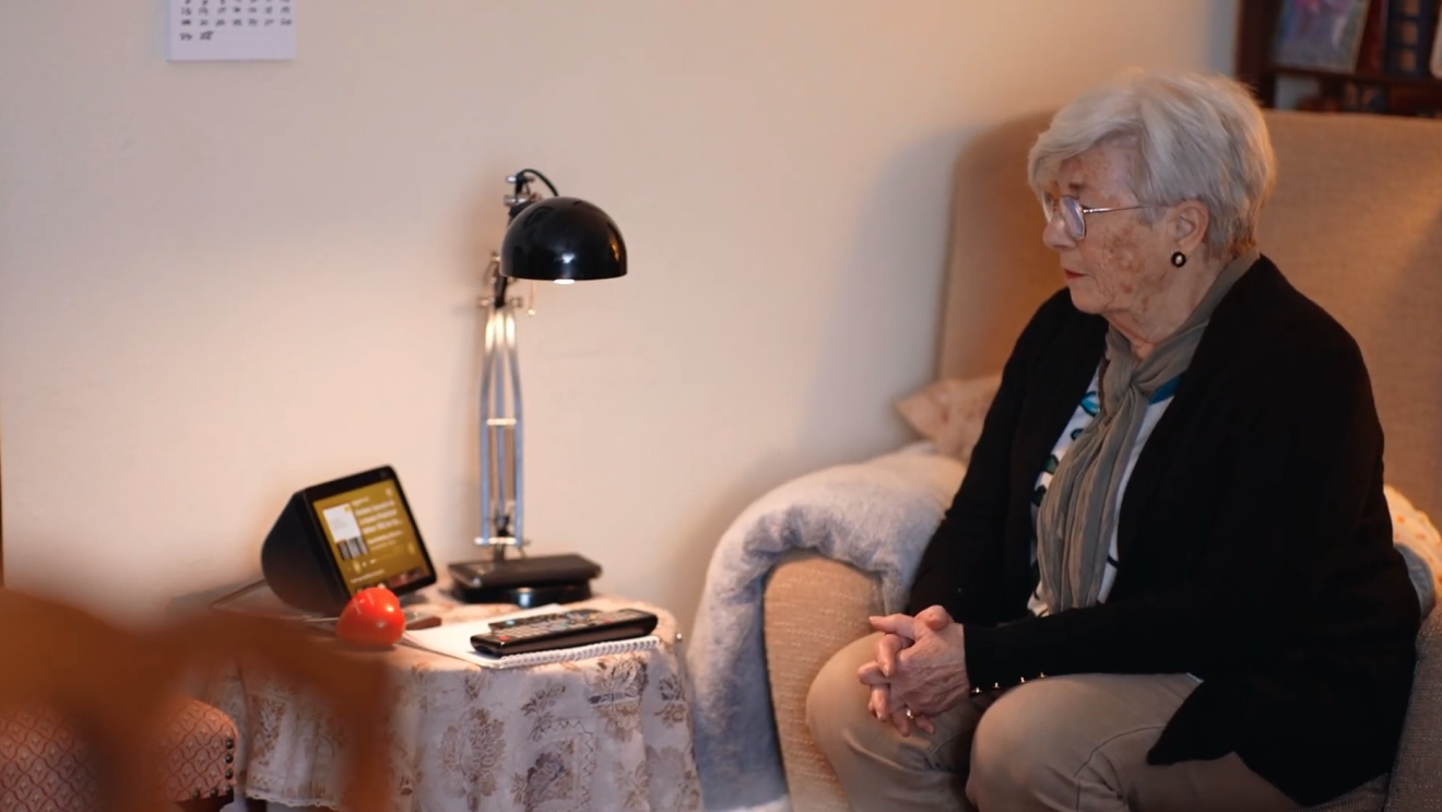 Una mujer mayor sentada en un sillón mirando a un dispositivo de Amazon con Alexa incorporado que está situado en una mesa auxiliar. 