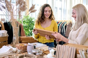 An image of a woman wearing a yellow shirt and another woman next to her wearing a cream shirt. They are in a store looking at a wooden product together. 