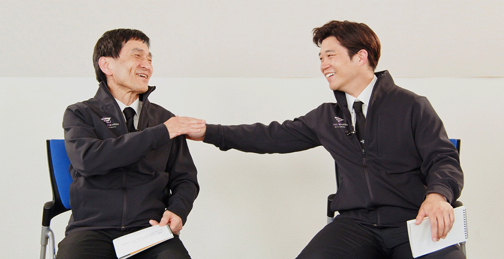 白い壁の室内で椅子に座った2人の人物が笑顔で談笑している。右側の男性が腕を伸ばして左側の男性の方に触れている。2人とも手に小さなノートを持っている。