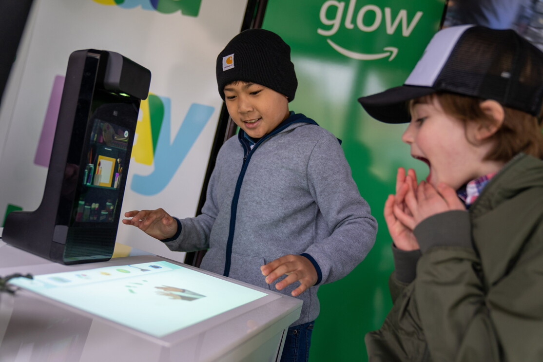 An image of two boys smiling while playing with the display version of Amazon Glow at an event. The device is projecting an interactive screen on a table and they are playing  the games and activities on it.