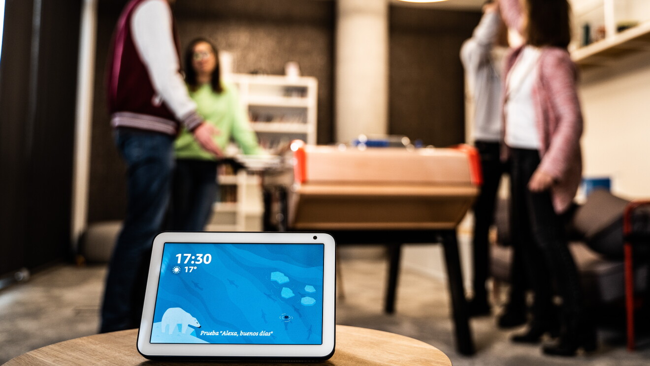 El dispositivo Echo Show en primer plano con la pantalla de color azul la hora y la temperatura. EL dispositivo se encuentra encima de una mesa. De fondo se ven cuatro personas jugando al futbolín. Dos chicos y dos chicas. Las parejas son mixtas y una de ellas se está celebrando que ha marcado un gol. 