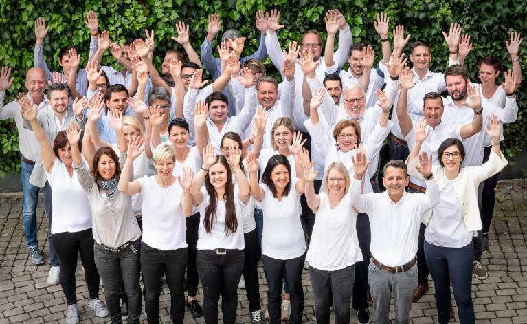 Das Team des Autozubehörproduzenten Walser aus Hohenems in Österreich steht in weißen T-Shirts bekleidet mit hoch gestreckten Armen auf einem begrünten Innenhof.