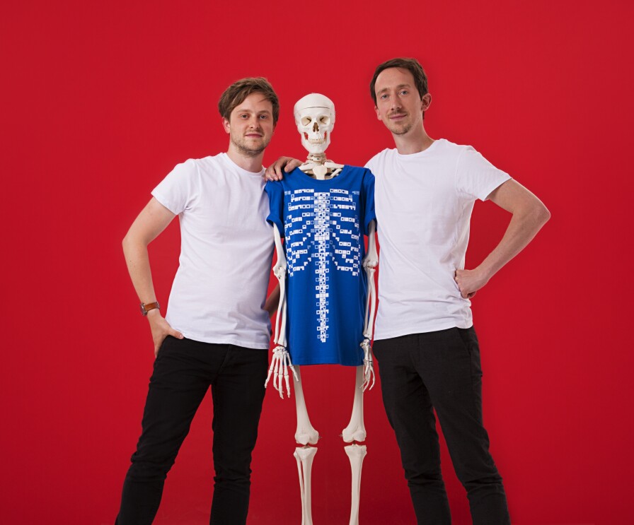 Ed Barton e Ben Kidd, fondatori di Curiscope. I due ragazzi, in t-shirt bianca e pantaloni nero, abbracciano uno scheletro con una t-shirt blu. Sfondo rosso. 