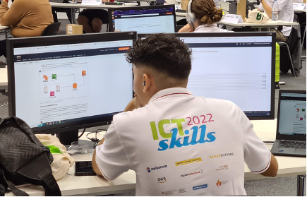 Ein Schweizer AWS Mitarbeiter vor 2 Computer-Bildschirmen. Man sieht ihn von hinten. Er trägt ein weißes T-Shirt auf dessen Rückseite "ICT 2022 skills" zu lesen ist.