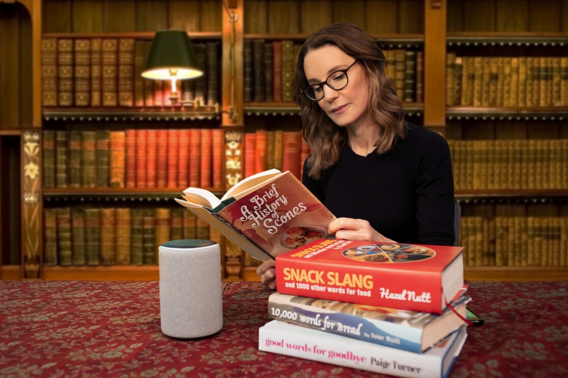 Susie Dent wearing glasses sat at a table, reading books about language and dialect