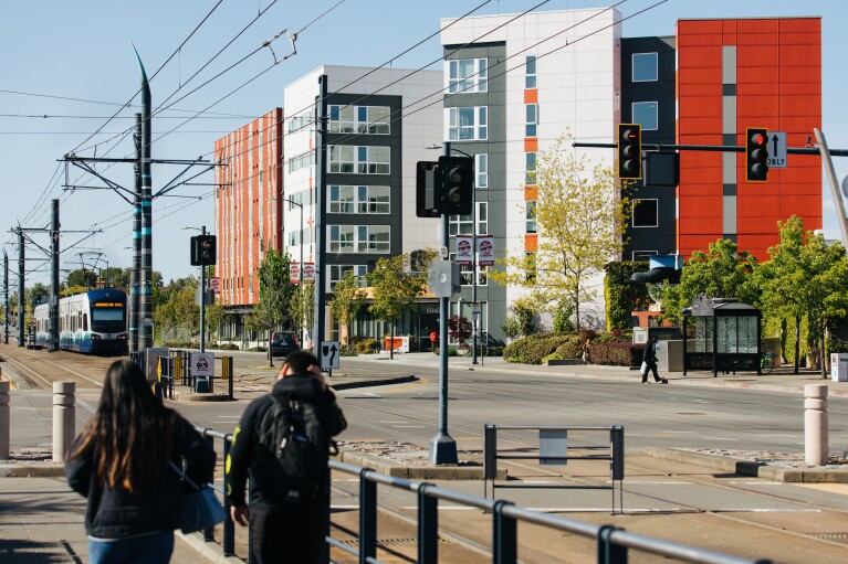 Two people dressed for cool weather walk along a sidewalk. Across the street are a number of condo buildings. A mass transit vehicle approaches the intersection.