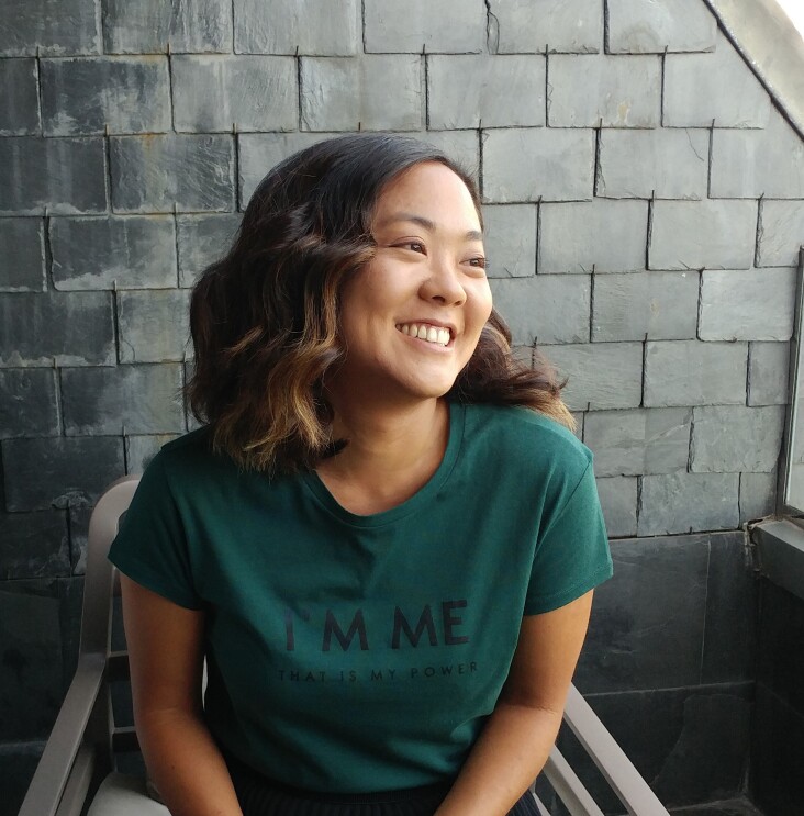 A photo of a woman smiling while looking away from the camera. She is standing in front of a brick wall wearing a green shirt.