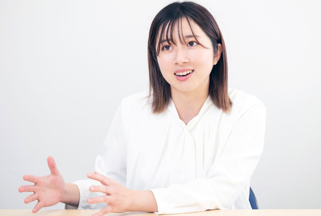 白い壁を背景に座った人物が、両手のジェスチャーを交えながら話している。白いブラウスを着用し、肩くらいの長さの髪。