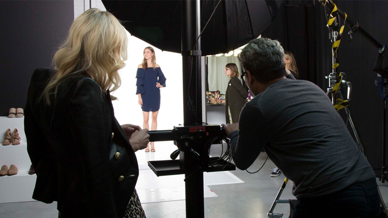 Two individuals, a male photographer and a woman in a leopard-print dress and dark jacket draped over her shoulders, photograph a model wearing a blue dress during an Amazon Fashion shoot.