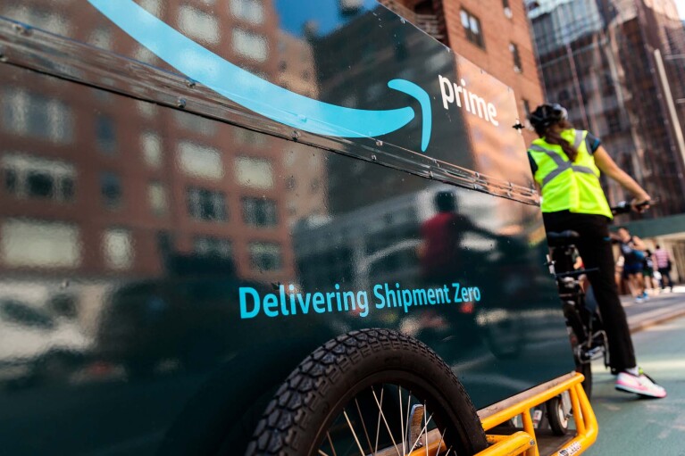 An Amazon delivery person riding a bicycle, with a bike trailer behind it. The trailer has "Delivering Shipment Zero" printed on it.