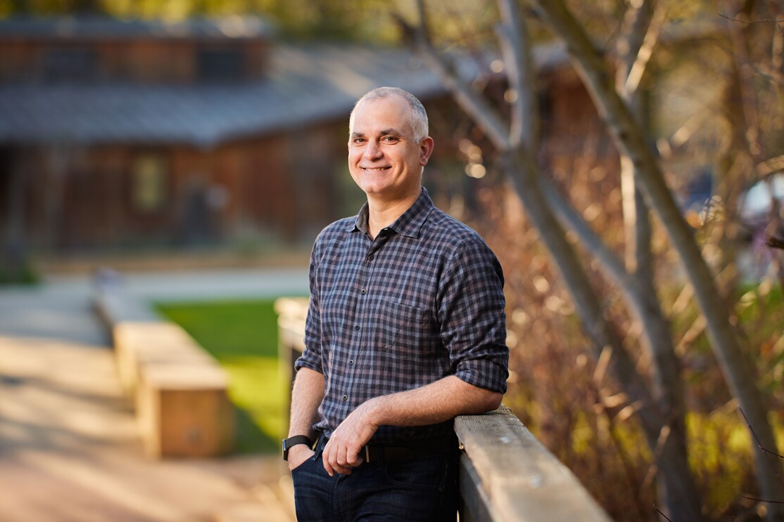 An image of John Ciancutti smiling for a photo in a park. He is leaning his left elbow on a railing and his right hand is in his pocket.