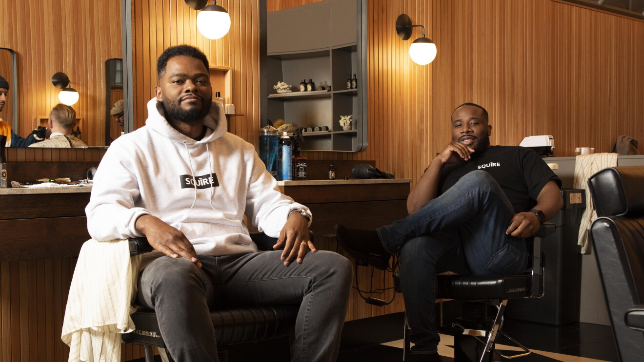 Two Black men, founders of the app Squire, sit in barber chairs in a barbershop setting. 