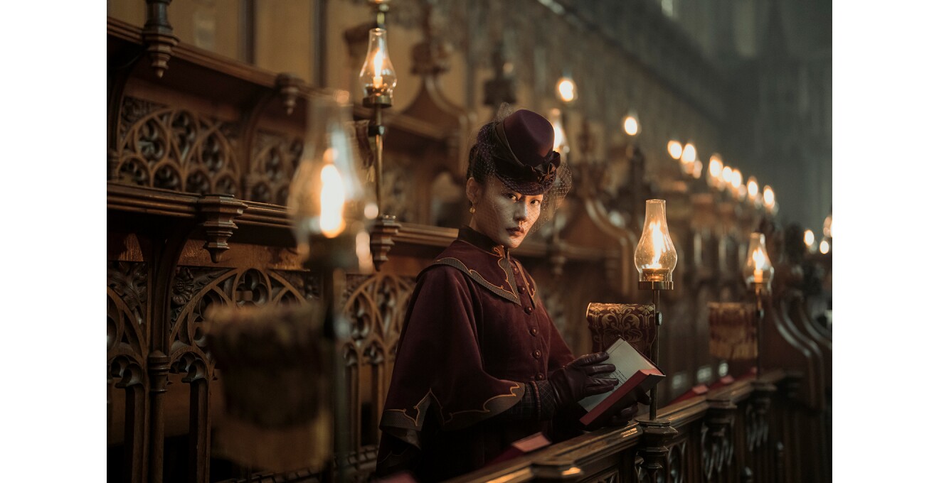 ゴシック風の教会の階段席に腰掛けて書物を開く女性。落ち着いた色合いの帽子とドレス、手袋で正装している。階段席には灯ったランプが並ぶ。